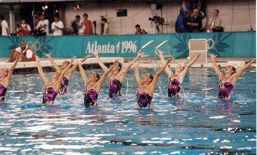 1996 US Olympic Synchronized Artistic Swimming Gold Medal performance. The team is in the pool jumping out of the water to their hips wearing puple costumes