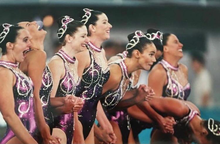 1996 Olympic Synchronized Artistic Swimming team seeing their perfect 10 scores come up on the scoreboard. The facial expressions are a whole range of emotions from mouth open smiles head flying back to crying.