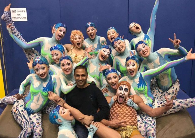 The Synchronized artisitc swimmers at the "O" Show from Cirque Du Soleil are in their brightly colored shades of blue and green head to toe bodysuit costumes. They are all piled together in the training room backstage taking a wild and funny photo with the resident physical therapist. They are making funny faces and is strage positions in a group.