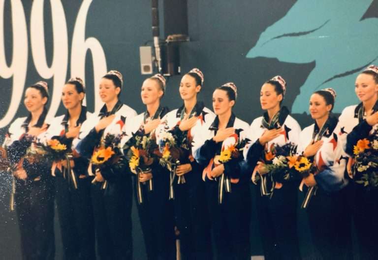 The 1996 Olympic Gold Medal Synchronized Artistic swimming team having received their gold medals are standing on the podium with their hands on their hearts, watching the United States flag raise and listening to the United States National Anthem play