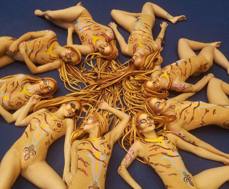 Synchronized swimmers in the O show in costumes tha are skin colored and wearing long wigs that look like rope. They are a variety of coppers colors. 9 girls are lying in a circle with their heads in the middle and their legs in artistic positions. 3 of these women are in perimenopause, but everyone looks like they are in their 20's.