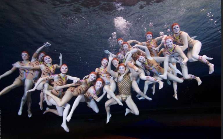 Picture of synchronized swimmers underwater posing all together at the "O" show by Cirque Du Soleil. They are floating and holding on to eachother, and very happy. Suzannah Bianco is in this picture.