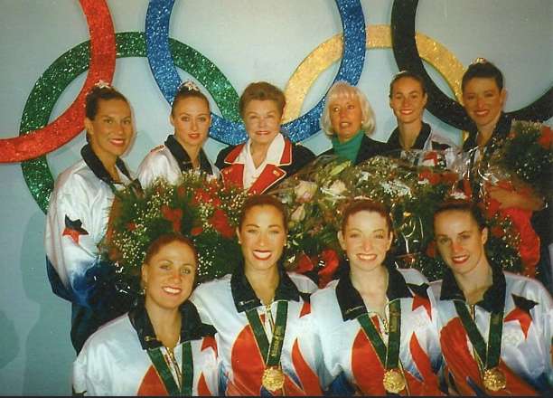 a photo of the 1996 synchronized artistic swimming gold medalists standing with Esther Williams, who is considered the mother of synchronized swimming with their gold medals and Olympic track suits on. Suzannah Bianco is part of this team