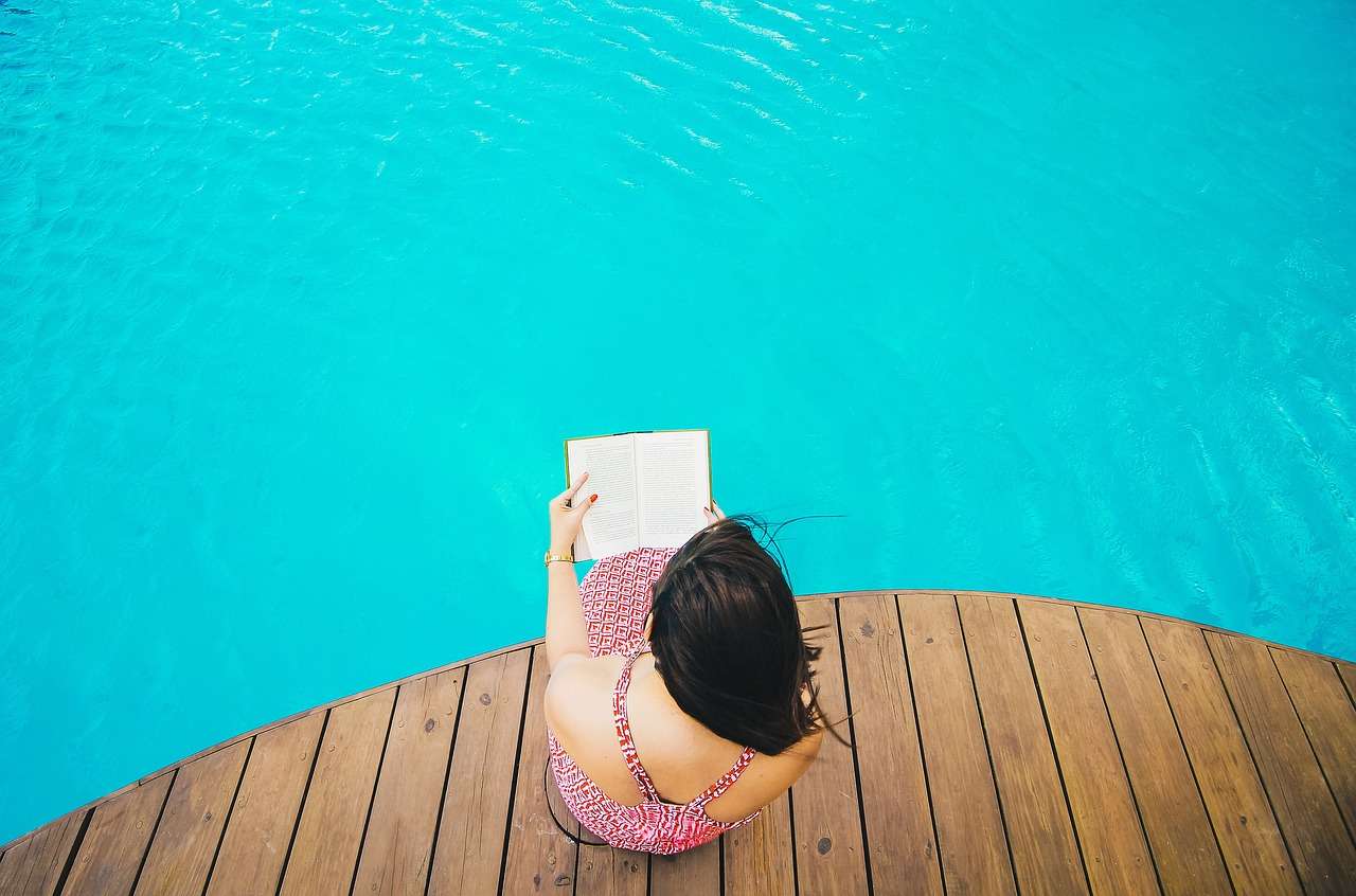 woman in red and white sundress, reading a book, sitting on a rounded wood plank deck over a blue body of water