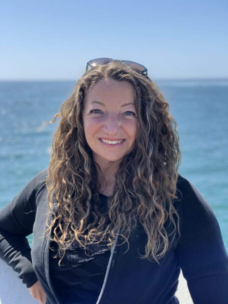A photo of Suzannah Bianco wearing a black hoodie and sunglasses on her head. she has long brown curly hair and the ocean is behind her.